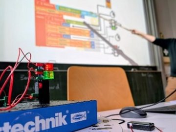 Eine Lehrkraft zeigt auf eine Präsentation an der Wand mit einem technischen Messgerät im Vordergrund.