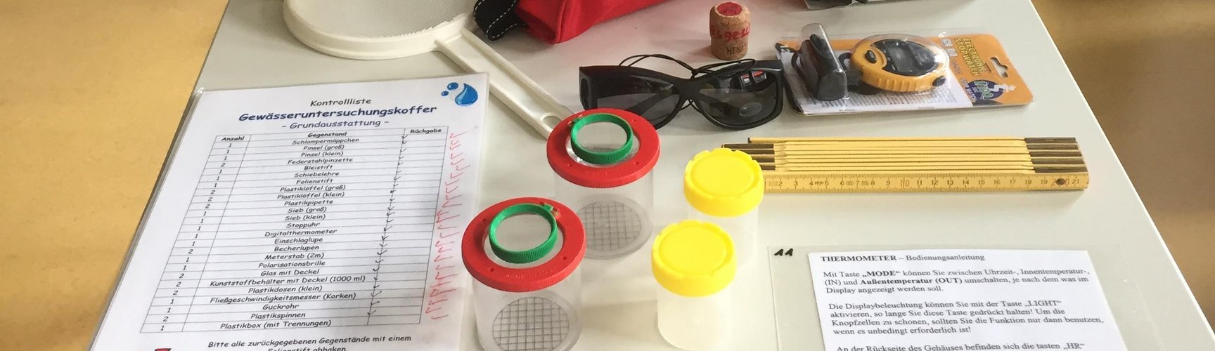 Auf einem Tisch steht ein Koffer mit der Beschriftung "Gewässeruntersuchungskoffer - Grundausstattung", davor liegen ein Sieb, Unterlagen, Schraubbecher, Sonnenbrille etc.