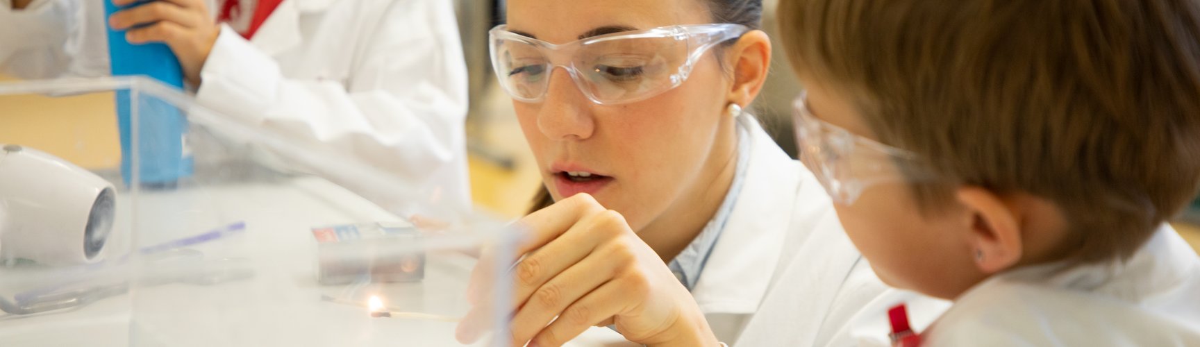 Eine Studentin macht zusammen mit einem Grundschulkind ein chemisches Experiment. Beide tragen Schutzbrillen.