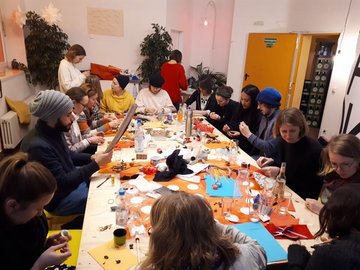 Eine Gruppe Studierender sitzt um einen Tisch und bastelt, auf dem Tisch liegen viele Materialien wie Papiere und Nüsse.