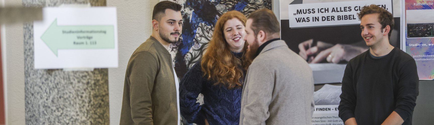 Vier Personen stehen an einem Stand des Faches Evangelische Theologie, im Hintergrund ein Aufsteller mit Plakaten.