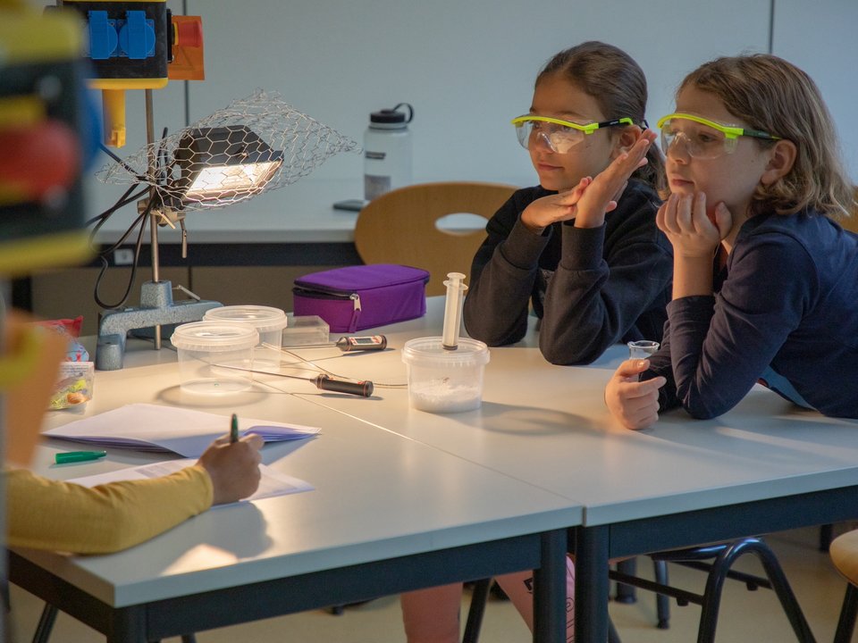 Zwei Grundschulkinder mit Schutzbrille verfolgen ein physikalisches Experiment