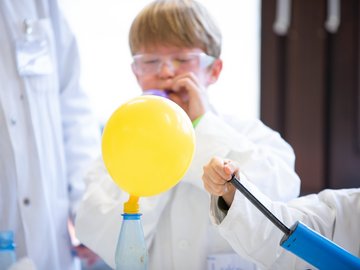 Ein Junge in Laborkittel und Schutzbrille bläst einen blauen Ballon auf. Vor ihm steht ein luftgefüllter gelber Ballon, der an einer Falsche befestigt ist.