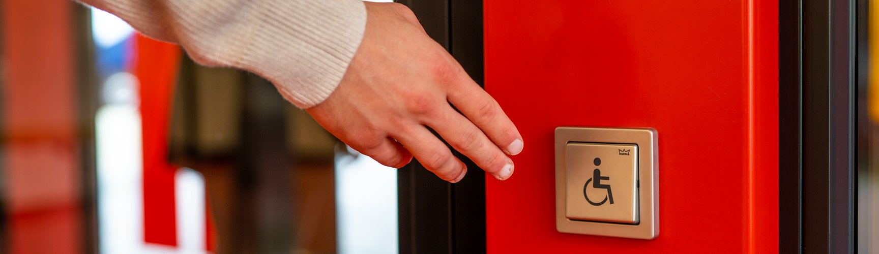 eine Hand ist dabei, auf einen automatischen Türöffner eines barrierefreien Eingangs zu drücken