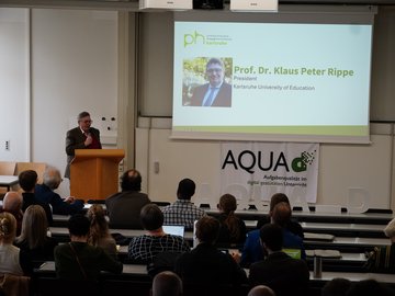Klaus Peter Rippe bei der Eröffnung der AQUA-d Konferenz 2025. Er steht am Pult vor einer Präsentation, im Vordergrund des Bildes sind Zuhörende in den Sitzreihen.