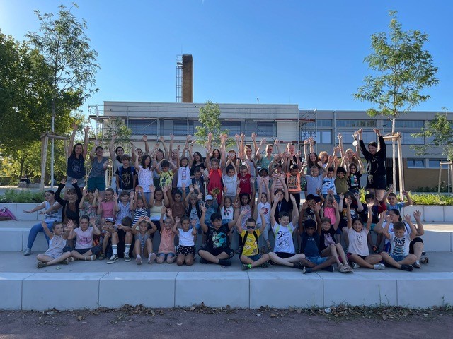 Vor einem Schulgebäude sitzen uns stehen Kinder in vier Reihen und heben jubelnd die Hände