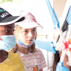 Drei Kinder mit Kaspe und Basecaps stehen für ein Experiment im Rahmen des Ferienprogramms RELAX vor einem kleinen Windkraftrad