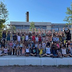 Vor einem Schulgebäude sitzen uns stehen Kinder in vier Reihen und heben jubelnd die Hände