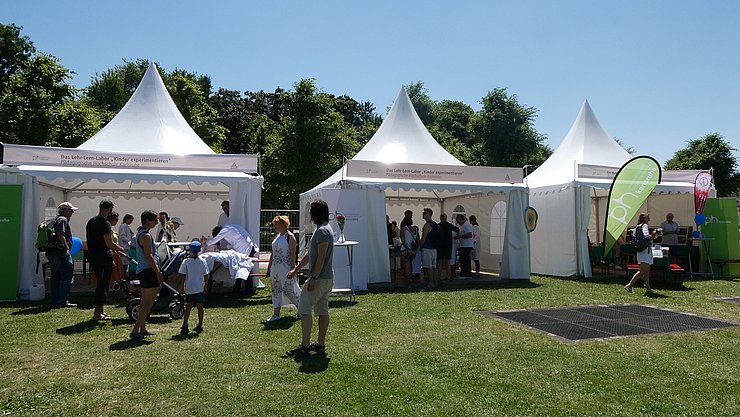 Drei Pagodenzelte der PHKA bei EFFEKTE 2023, im Hintergrund Bäume , vorne Wiese, in und vor den Zelten sind Menschen unterwegs