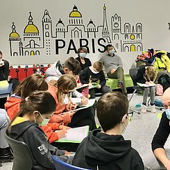 In einem hellen Gruam der Brettener Gesamtschule sitzen Kinder im Kreis und diskutieren mit einem PH Studenten über Philosophie. auf der wand im Hintetrgrund ist schematisch ein Häuserzug mit dem Schriftzug "Paris" zu sehen.