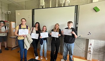 Zertifikate EntreCo Think Hackathons Fünf jugendliche Schüler:innen zeigen stolz ihre durch einen Hackathon des Projekts EntreCo Think erworbenen Zertifikate vor einem Whiteboard.