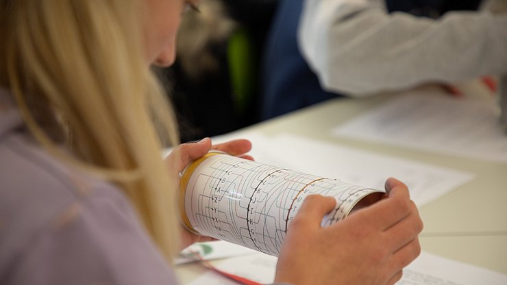 LLL_Informatik_Abstraktion Eien Schülerin von hinten, die konzentriert auf bildlich dargestellte Programmierschritte auf einer Rolle blickt.