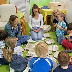 Kinder sitzen mit zwei Jungen Frauen auf dem Boden und sortieren Plastikeier in Eierschachteln