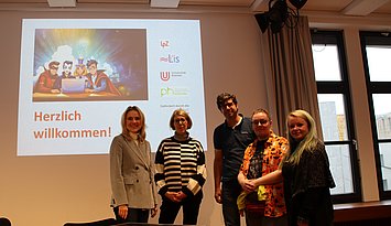 2024_Fanfiction_Schreibwerkstatt Dei Projketpartner, vier Frauen und ein Mann, stehen vor dem Titelbild der PPT für den Kick-Off des Projekts in einem Seminarraum.