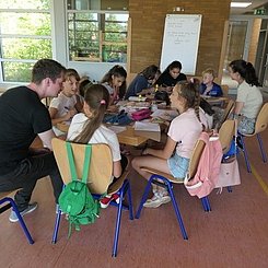 In einem Klassenzimmer sitzen acht Kinder an einem großen Tisch, ein Student sitzt erklärend dabei