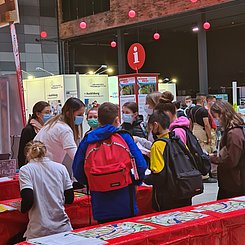 Beim Blick in die Halle der Science Days sieht man Kinder von hinten an einem Tisch mit roter Bedeckung und Utensilien für mathematische Experimente, wie sie sich von einer Studentin etwas erklären lassen.